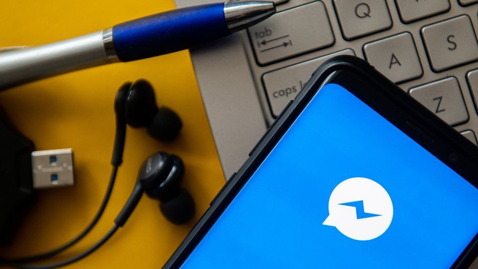 The Facebook messenger logo is seen on a mobile phone lying on a laptop, next to some pens and earphones and a USB cable