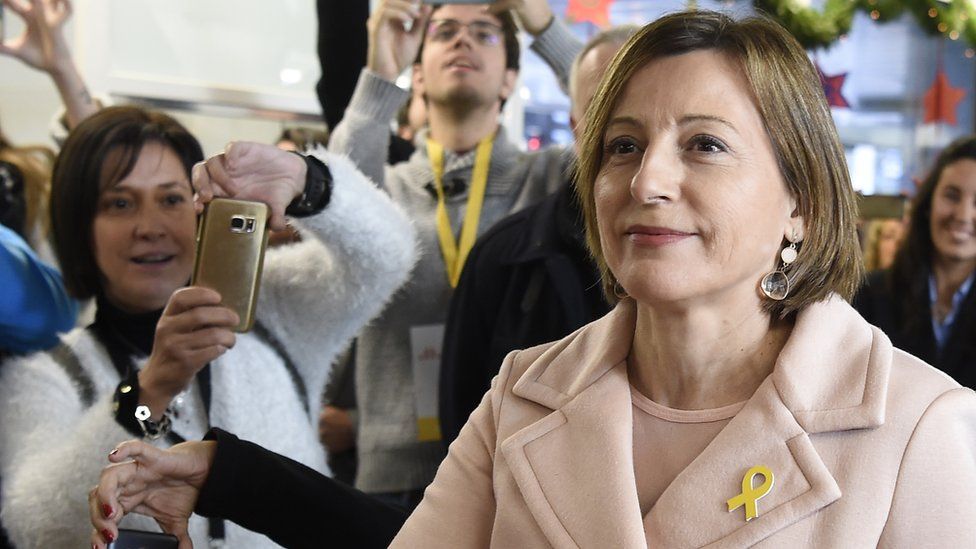 Carme Forcadell looks up and into the distance as bystanders photograph her - her right arm is extended, out of frame, towards the ballot box, and on her lapel she wears the yellow ribbon adopted as a symbol of solidarity with other jailed leaders