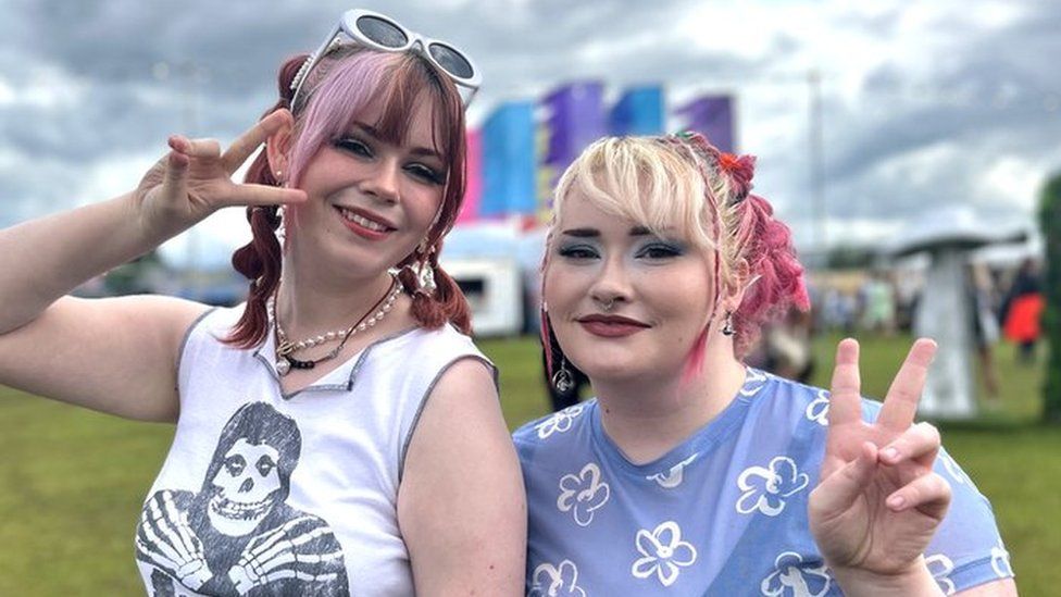 Yasi and Emily pose with peace signs at 90s Baby Festival, they're smiling. Yasi is wearing a white t-shirt and Emily, a baby blue sheer top. They both have pink highlights in their hair.