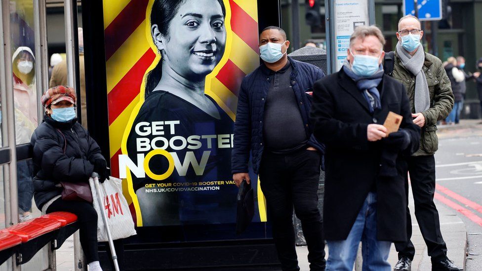 People wearing face coverings to combat the spread of the coronavirus wait at a bus stop displaying a government advertisement promoting the NHS Covid-19 vaccine Booster program in London on December 17, 2021.