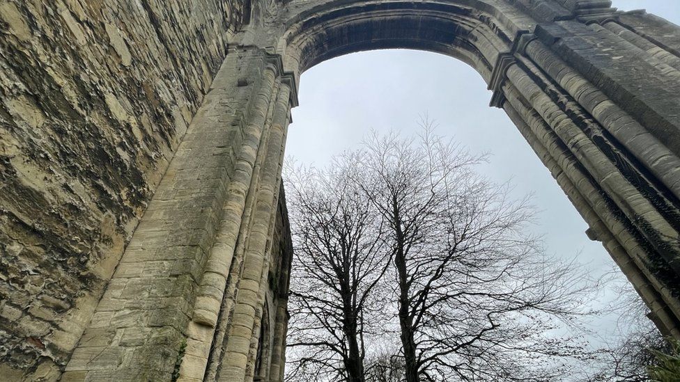 A large medieval archway as a ruin, with a grey sky and trees