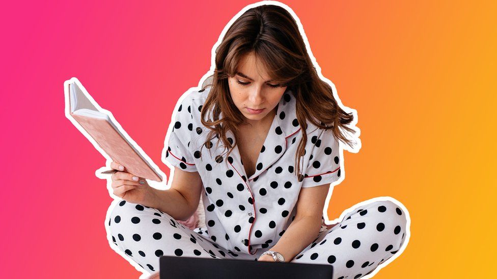 A woman in her pyjamas holds a notebook while reading from a laptop, cut out against a pink and orange gradient