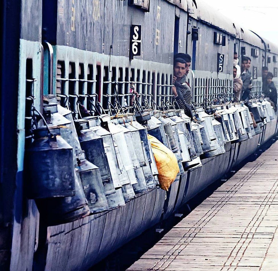 Milk cans are tied to the outside of a train as milkmen peep out of the doors.