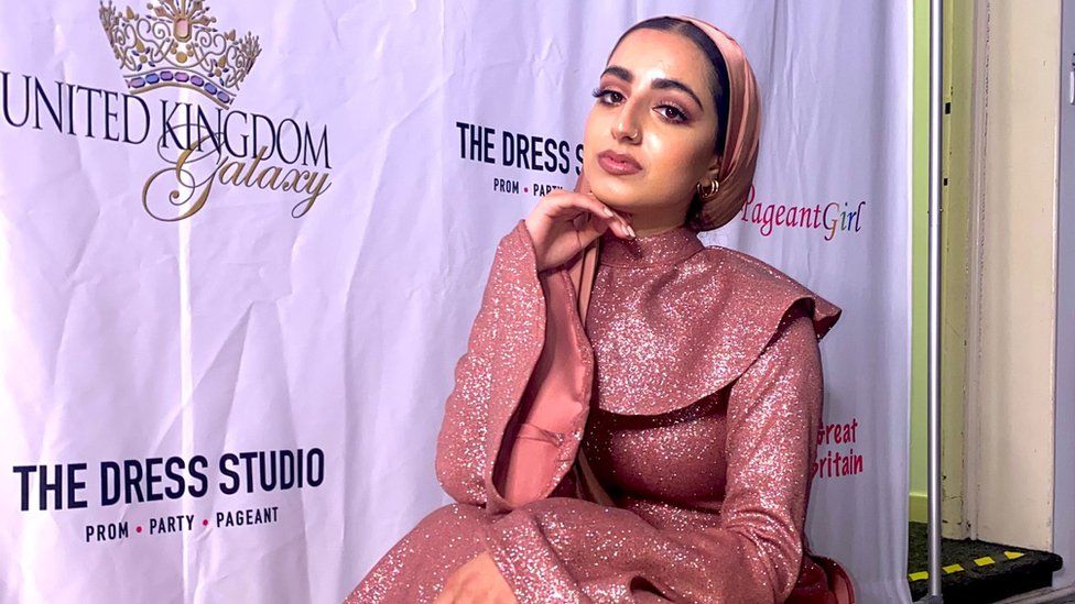 Fatima is wearing an glamourous pink long sleeved dress and matching hijab. She has a nose ringe piercing and has her right hand under her chin