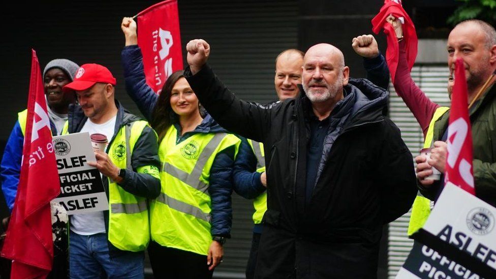 Bearded man wearing large blue coat stands with one arm raised among strikers wearing yellow hi-viz