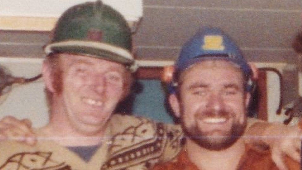 Two men in hard hats smile at the camera