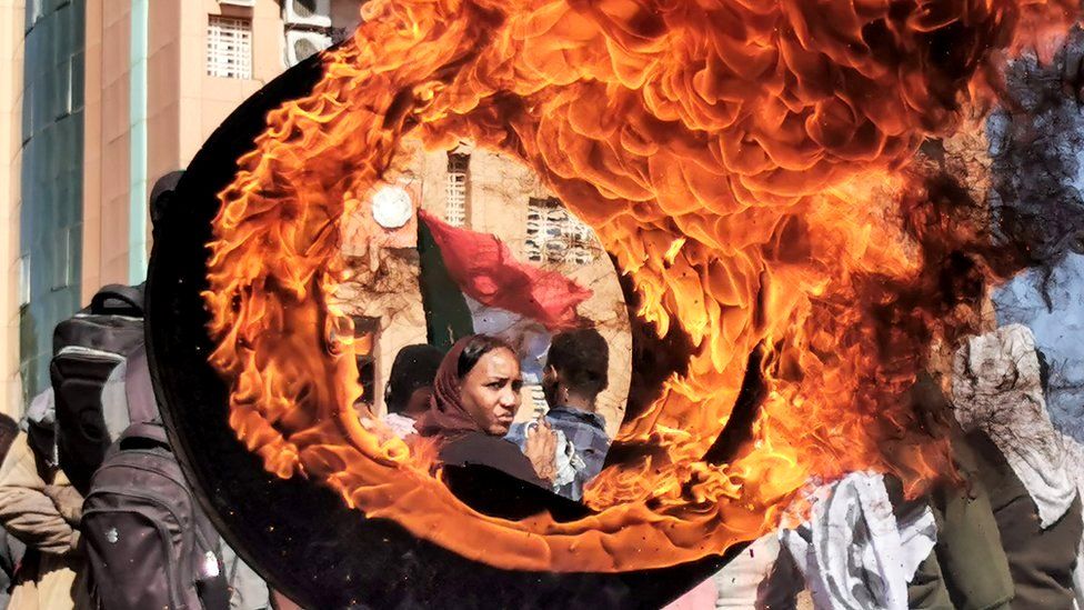 Демонстрант смотрит из-за пылающей шины на импровизированную баррикаду, воздвигнутую во время протеста с требованием гражданского правления в городе-побратиме суданской столицы Омдурмане, 4 января 2022 г.