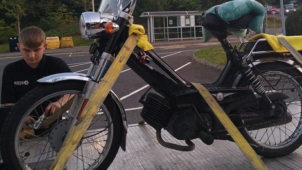 A black moped strapped to a recovery truck with yellow straps