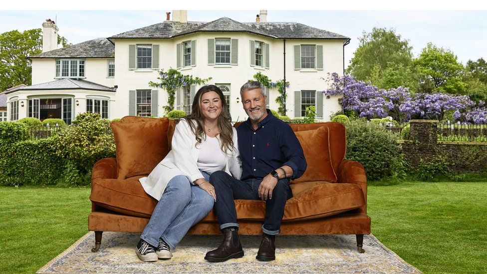 Jess and her dad Roger who are on new dating show My Mum, Your Dad pose in front of the country house where the show is set. They are sat on a brown sofa which is on a large rug on a green grass lawn and behind them are some stone steps and bushes, and then a grand white country house where the show is set. Jess is wearing a white top and white blazer with blue jeans and navy Converse shoes. She has her brunette hair curled in a wavy style and it is worn down over her shoulders. Roger is wearing a navy shirt with navy jeans and dark brown boots and his greying hair is styled up over the side of his head and he has a greying beard too. Both are smiling for the camera.