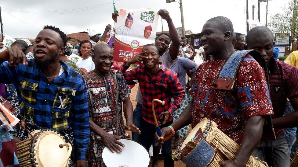 Nigerian men on the streets celebrating with talking drums and holding up election poster.