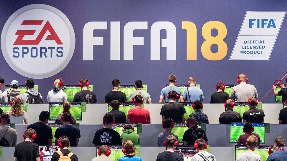 Many people are facing away from the camera playing the Fifa 18 video game. Above them, a sign reads Fifa 18.