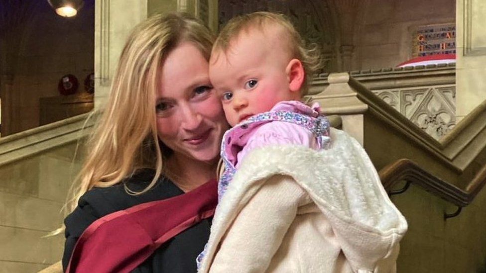 Charlotte in her graduation robes on the university steps holding her daughter Maya