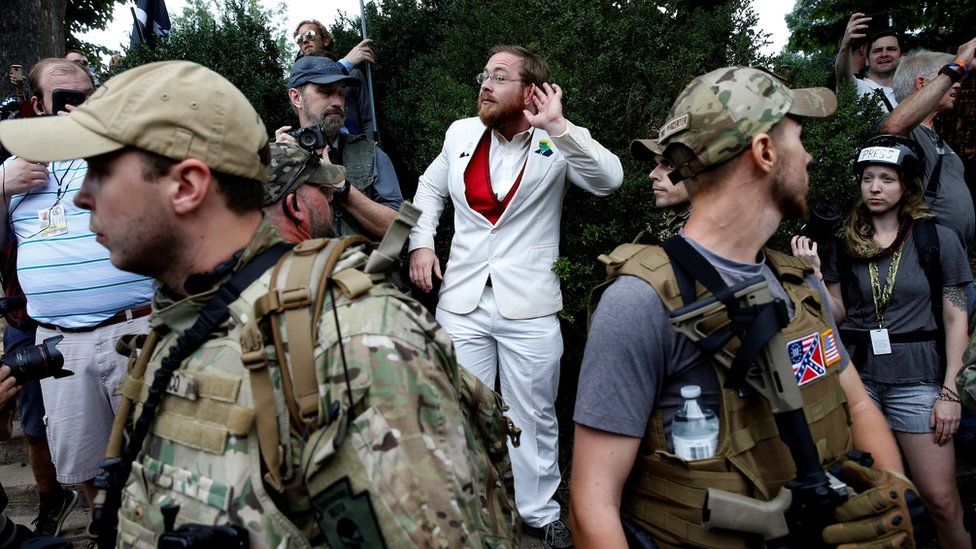 man in white suit standing behind two heavily armed men in bulletproof vests