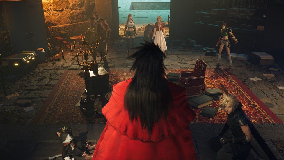 A man with long black hair and red velvet cloak is seen from behind, looking down at six other characters from an elevated position. They are in a candlelit room that looks Medieval in style, with ornate furniture, an opulent rug and stone floor.
