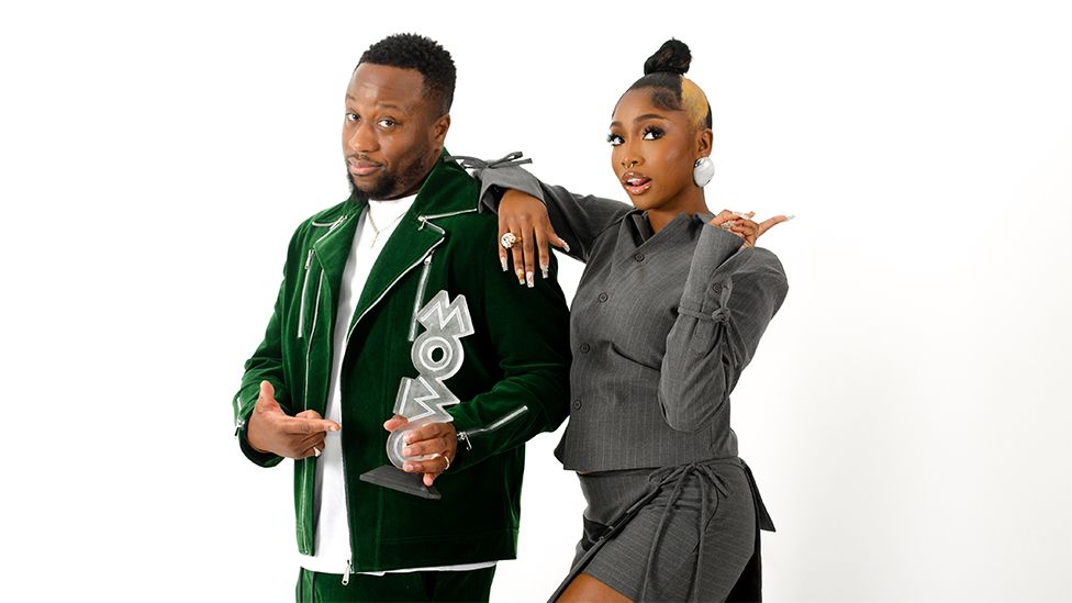Babatunde and Indiyah on a white background looking at the camera. Babatunde on the left is wearing a white tshirt under a green blazer, holding a silver award which spells out "MOBO". Indiyah on the right has her right arm over his shoulder, and is wearing a grey outfit with silver earrings. Her left hand is doing a "peace" sign.