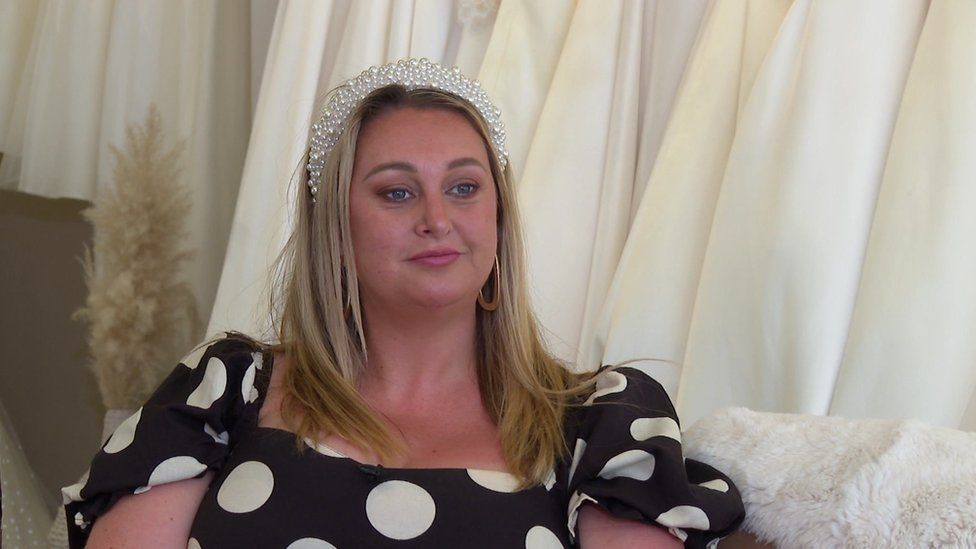 A woman with long, blonde hair wearing a black and white spotty dress, sat in her bridal shop.
