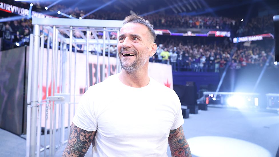 CM Punk, a white man, at Survivor Series, wearing a white tshirt with his arm tattoos visible. He is looking at the crowd, with the background full of fans and flashing white lights.