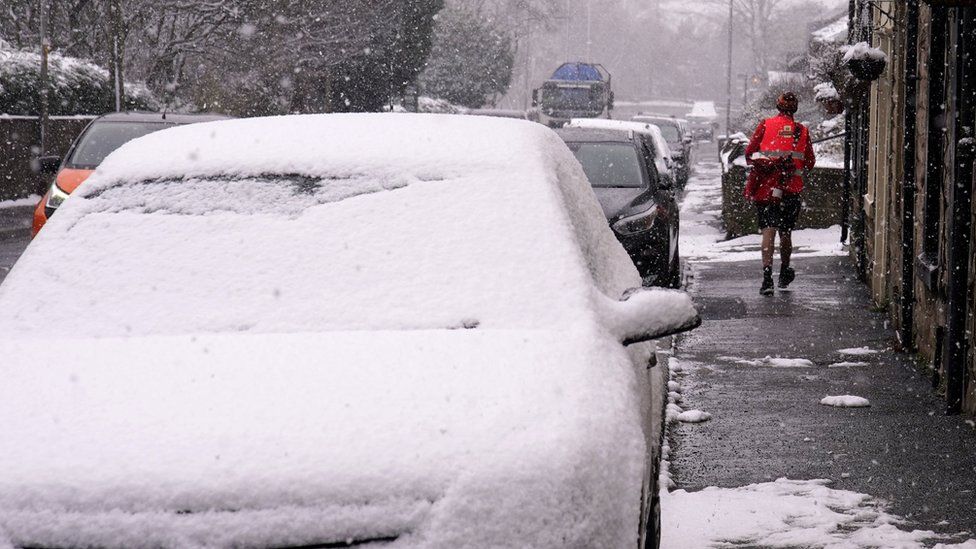 A postal delivery worker wearing shorts in heavy snow fall during his delivery rounds in Oldham, Greater Manchester