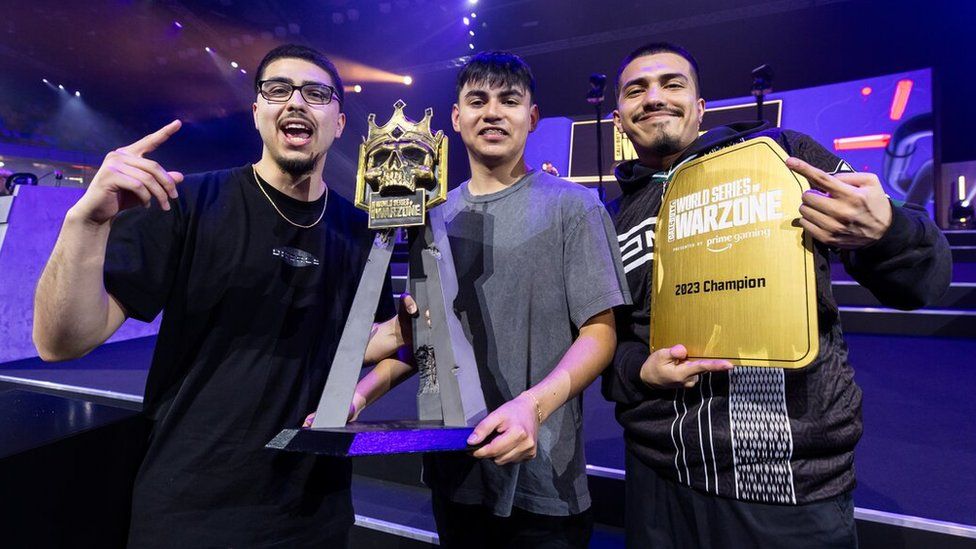 Three young men celebrate winning the World Series of Warzone global final. They show off the competition trophy - a golden skull, with a crown, on a tripod. One of the men also holds a gold plaque with "World Series of Warzone: 2023 Champion" written on it.