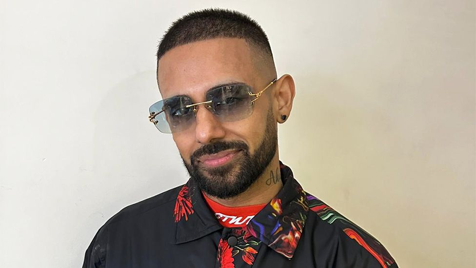 Jaz, a man, wearing sunglasses and smiling at the camera. The background is plain white. He is wearing a black shirt with flowery collar.