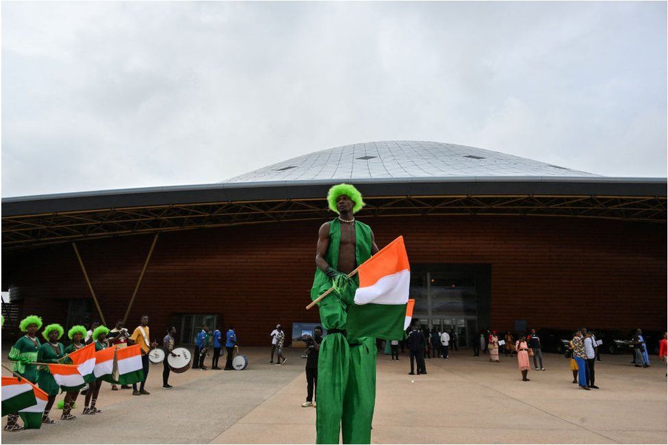 Man in stilts wearing Ivorian flag colours and a green wig.