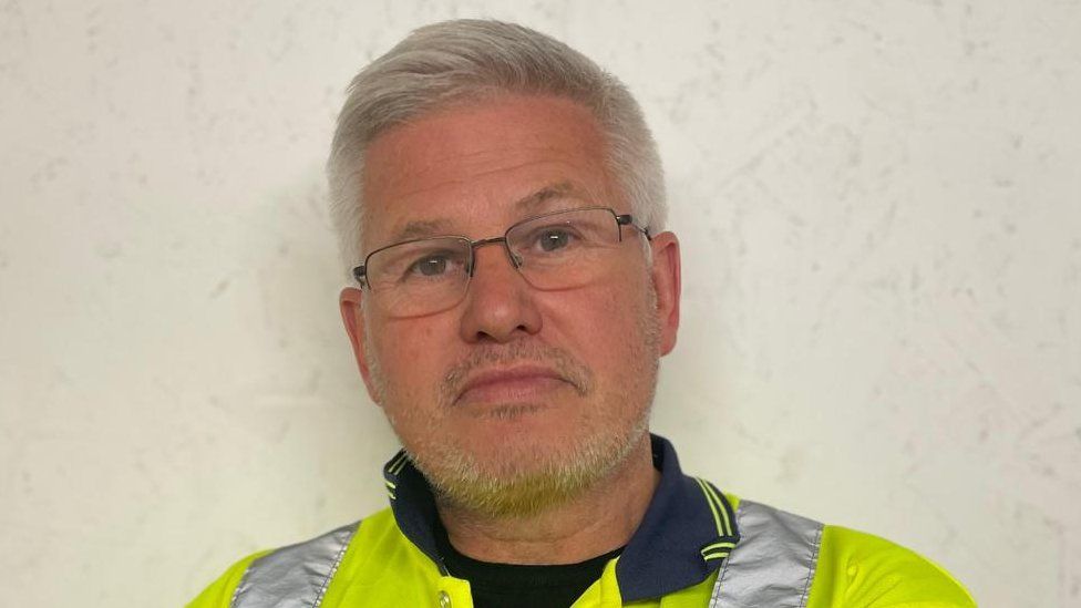 Landlord Andrew McCausland stands in a high vis jacket arms folded. He has silver cropped hair and light stubble. He's wearing glasses and looking at the camera.