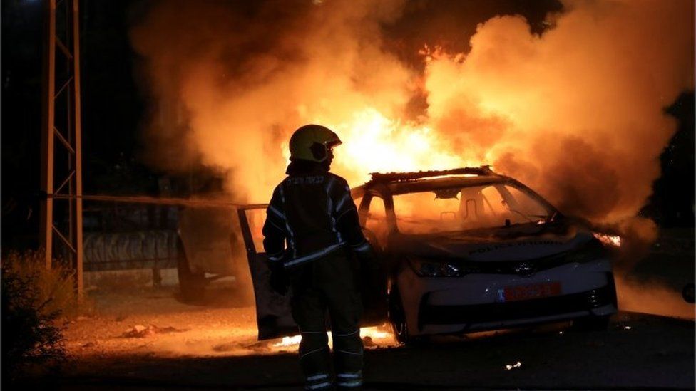 Израильский пожарный стоит возле горящей израильской полицейской машины во время столкновений между израильской полицией и представителями арабского меньшинства страны в арабо-еврейском городе Лод, Израиль, 12 мая 2021 года.