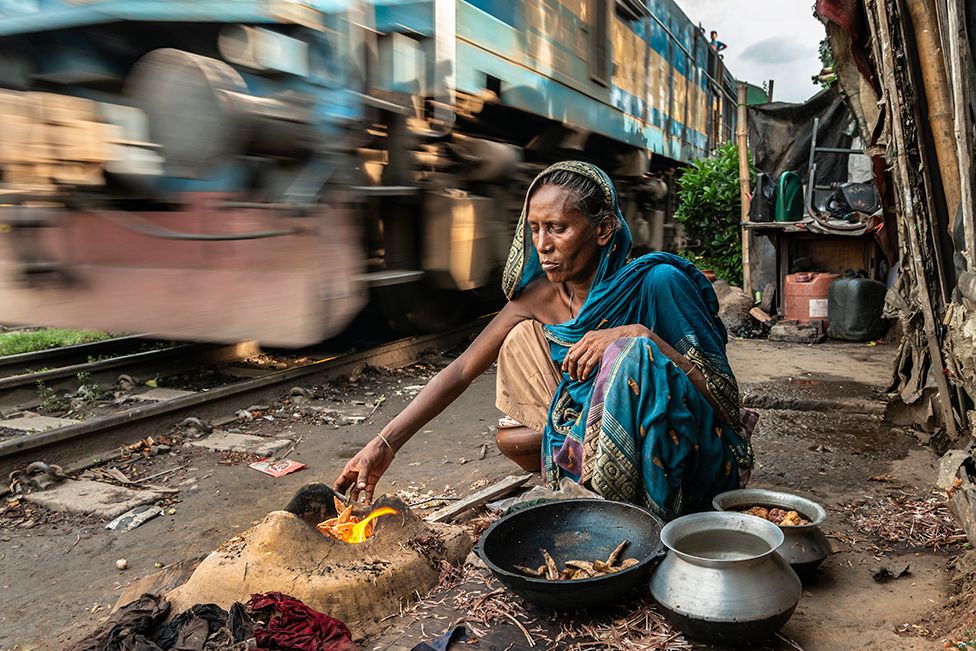 A woman cooks a meal over a fire as a train hurtles by behind her