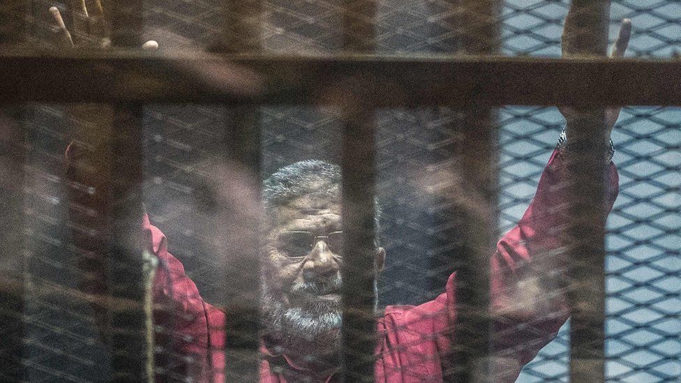 Mohammed Morsi gestures from behind bars during a trial in Cairo on 23 April 2016
