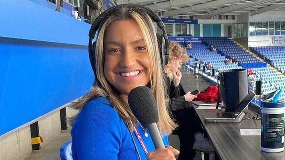 Commentator Millie Sian in the commentary position at Leicester City's King Power Stadium holding a microphone and wearing a blue top
