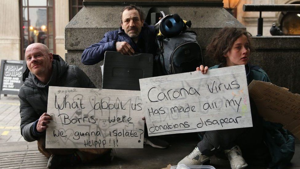Homeless people hold signs reading "What about us Boris - were we gunna isolate? Help!", and "Corona Virus has made all my donations disapear"
