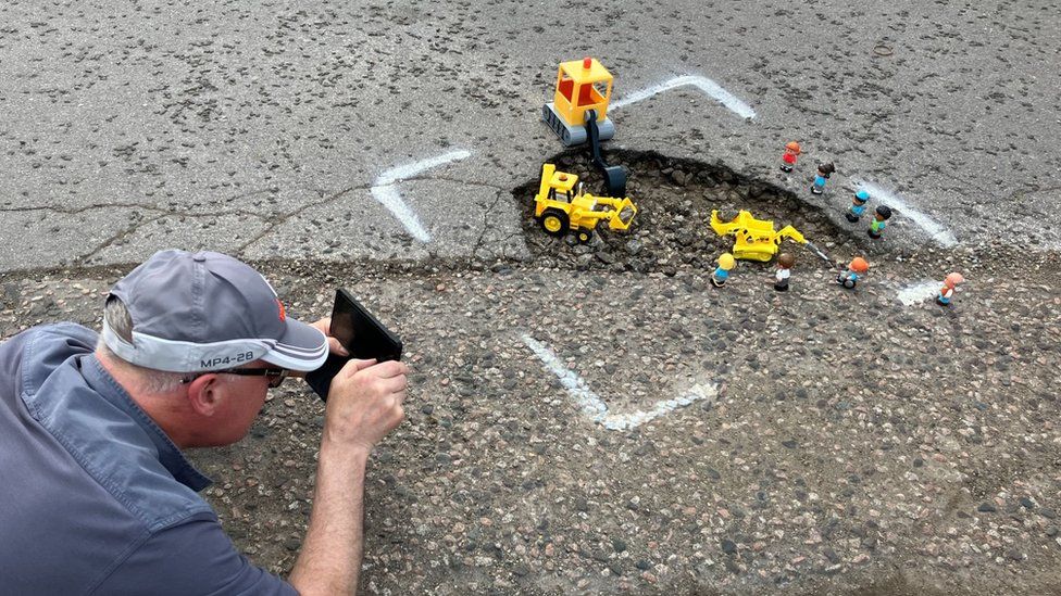 A man taking photos of pothole with toy diggers in it