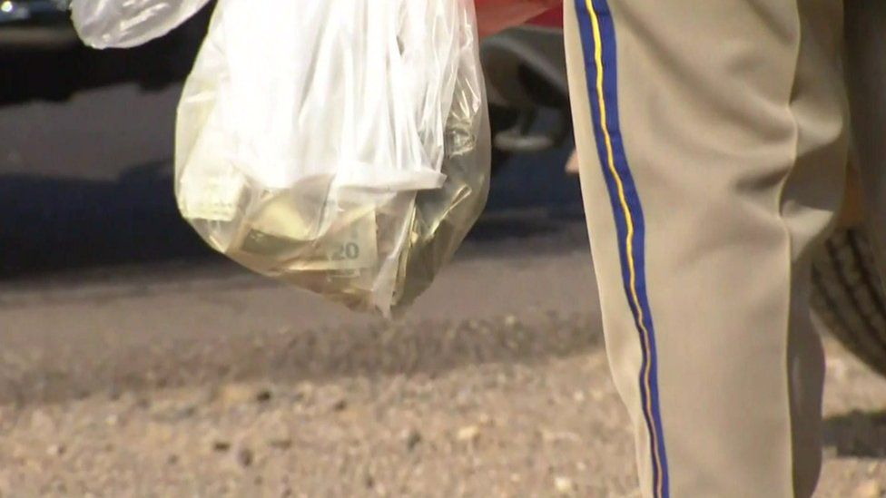 A police officer holds a carrier bag filled full of US dollars