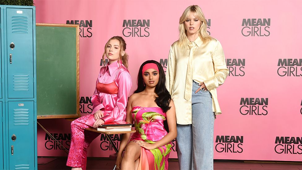 Bebe Wood, Avantika and Reneé Rapp stood in front of blue school lockers and a pink Mean Girls photoboard