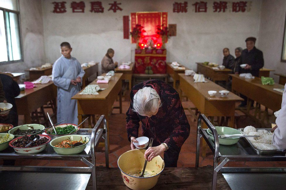 An elderly Chinese resident serves herself food in the food hall of the Ji Xiang Temple and nursing home in Sha County, Fujian province, China, on 18 March 2016. Months and other residents can be seen in the background, at long dining tables.