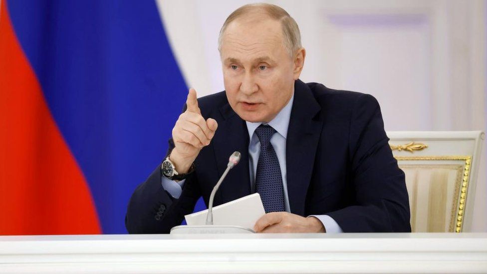 Vladimir Putin points as he speaks into a microphone from an office in the Kremlin