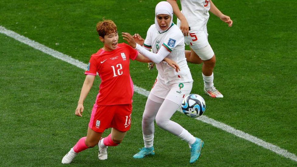 South Korea's Moon Mi-ra and Morocco's Nouhaila Benzina battle for the ball in their Women's World Cup game. Mi-ra is wearing a red football shirt and shorts that both have a pink trim and pink socks with white football boots. Her shirt has the number 12 on the front. She has short hair and is shouting out. Benzina is wearing an all white football kit and her shirt has the number 3 on the front. She has a white hijab (headscard) on her head. She also has white leggings on under her shorts and is wearing light blue football boots. Both players are looking at the white football which is to the right side of Benzina.
