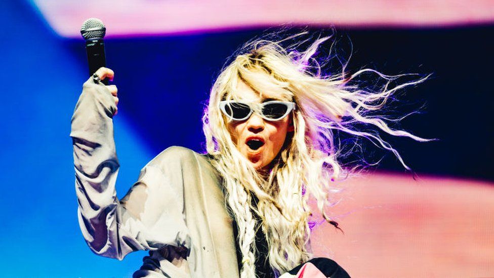 Grimes performing at Coachella. She is holding a microphone, and wearing sunglasses with her mouth open, her blonde hair is blowing in the wind.