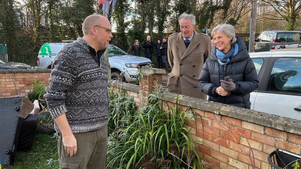 The duchess and a man in a brown coat speaking over a garden wall to Alistair Sinclair