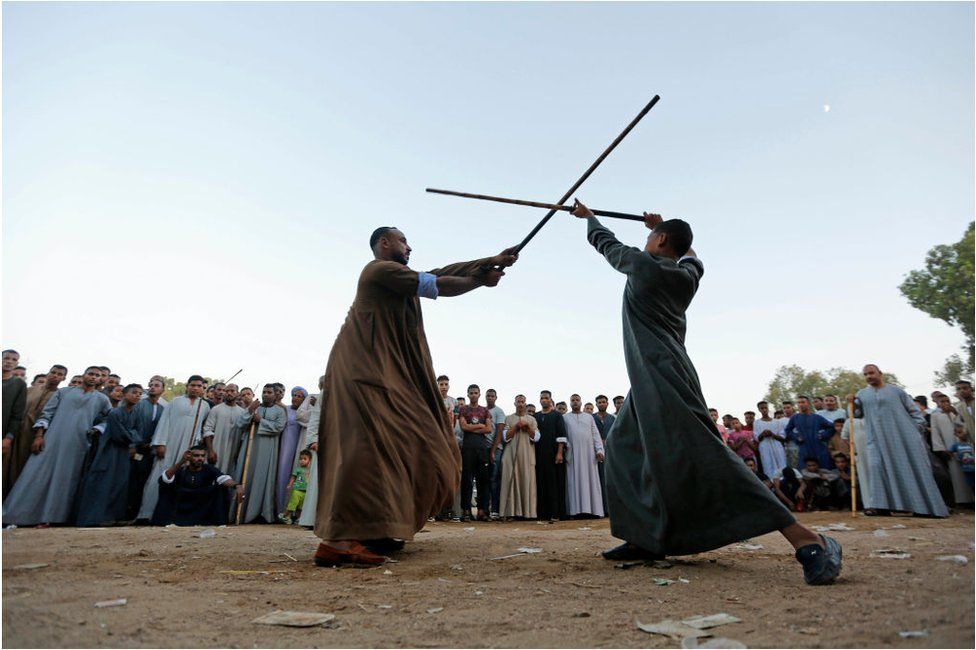 Two men with spears clashing against each other. They are surrounded by a crowd of people looking at them compete. They are outside, there are blue skies.