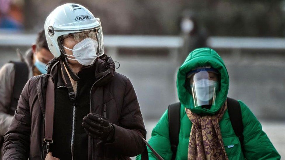 A man wearing a helmet and n95 face mask walks through the streets and a woman wearing a mask and faceshield behind him