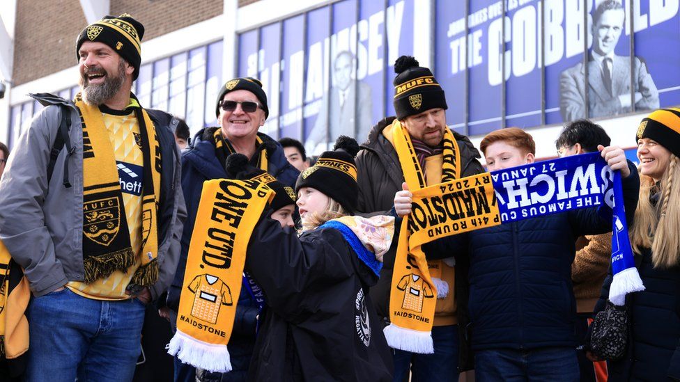 A group of Maidstone fans wearing yellow and black Maidstone United kits, hats and scarves gather outside the Ipswich stadium