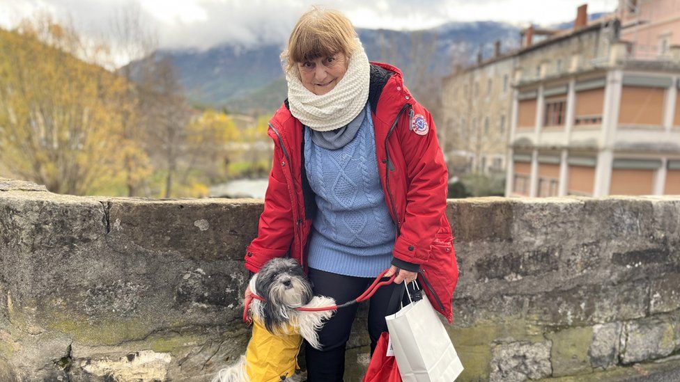 Martine Vincent, a resident of the French town of Quillan, seen with her pet dog. She is wearing a red coat and standing on a stone bridge. There are mountains in the background.