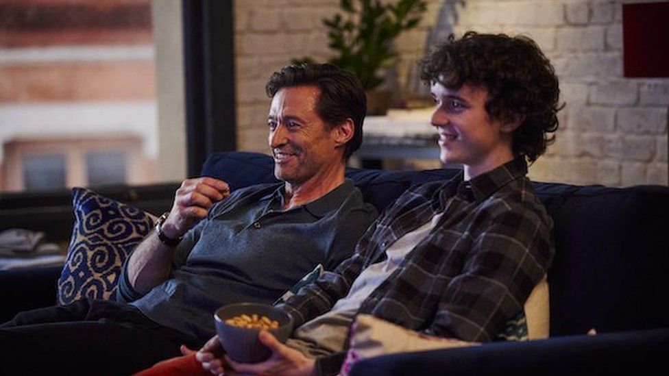 Hugh Jackman and Zen McGrath sit on a sofa together, smiling.