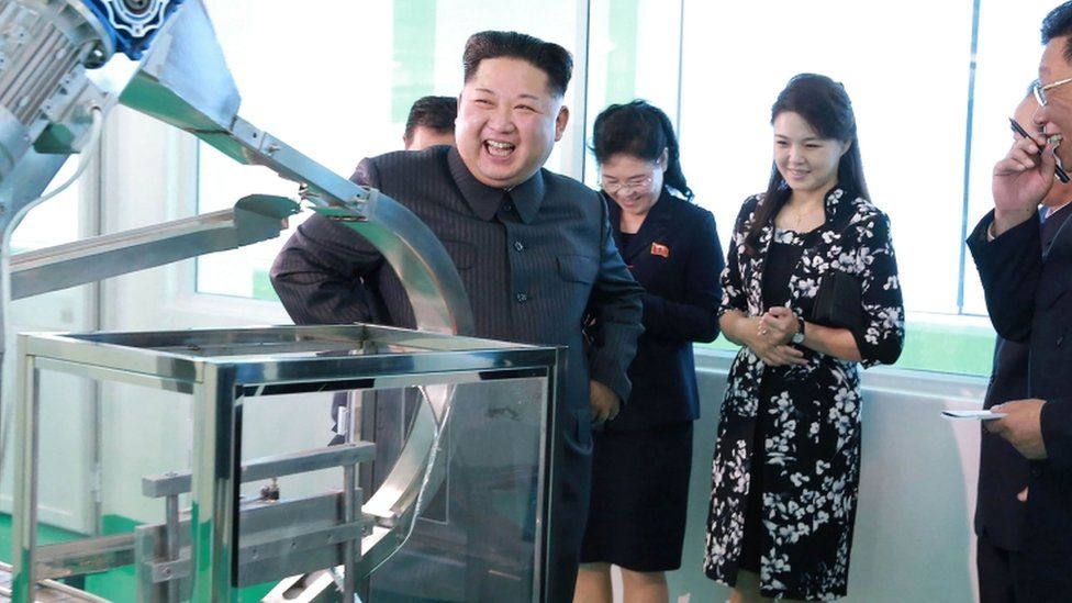 Kim Jong Un smiles toward electronic device in newly renovated facility with senior officials and his wife in the background