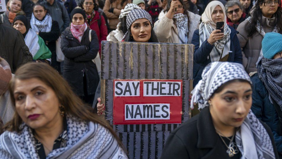 A woman in a crowd with a placard saying "say their names"