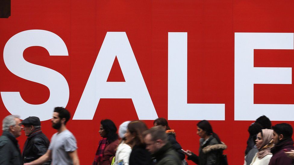 Large Sale sign in the window of a shop as people walk past outside