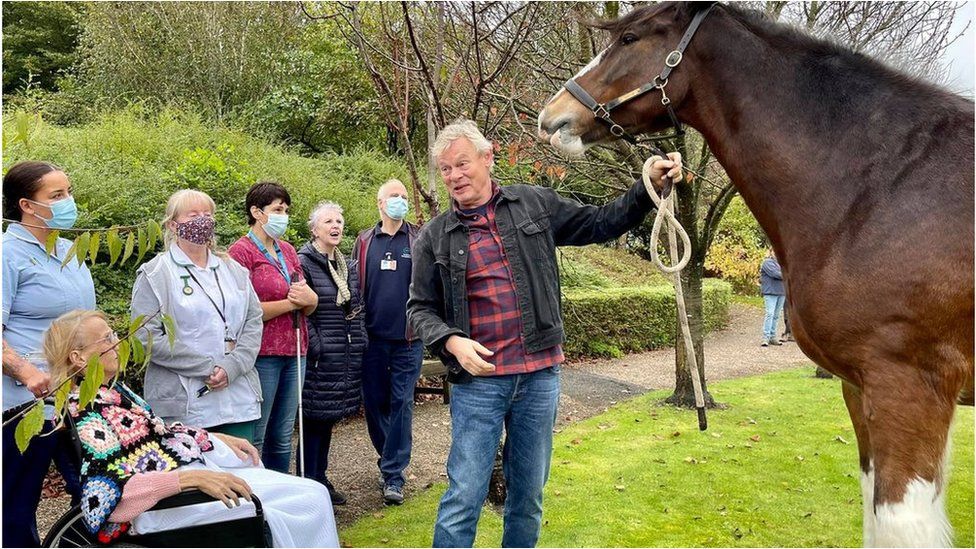 Martin Clunes holding the reins of a large brown horse surrounded by patients and staff