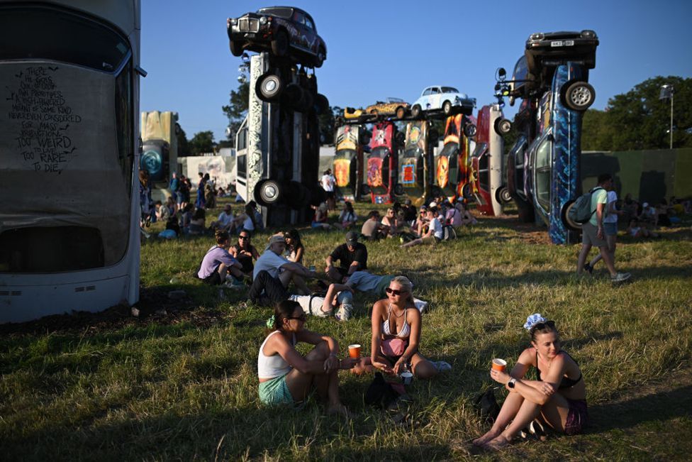 Festival-goers rest in the sunshine by the Carhenge art installation, which is composed of painted vehicles piled on to of each other, on the opening day of the Glastonbury festival at Worthy Farm in the village of Pilton in Somerset, southwest England, on June 26, 2024. 