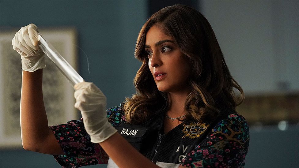 An image of Mandeep from the TV show CSI: Vegas, in which she is wearing gloves, a police vest and is looking at a piece of white tape.